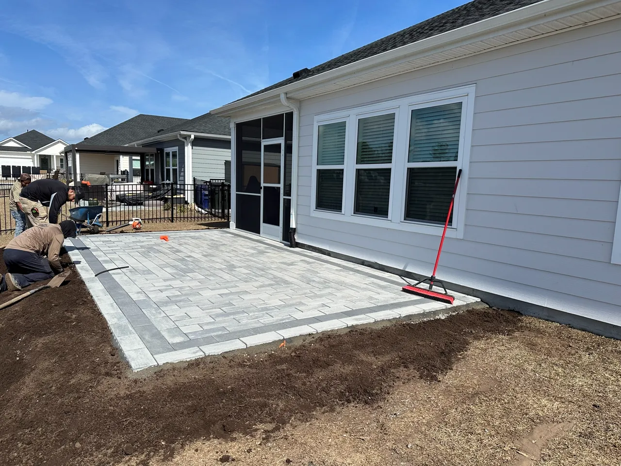 Crew laying pavers on a fresh base beside a house, mid-build.