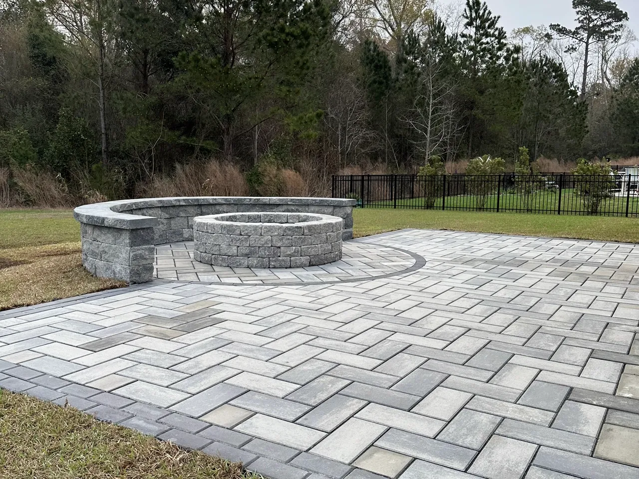 Wide view of a paver patio and stone fire pit against a tree line.