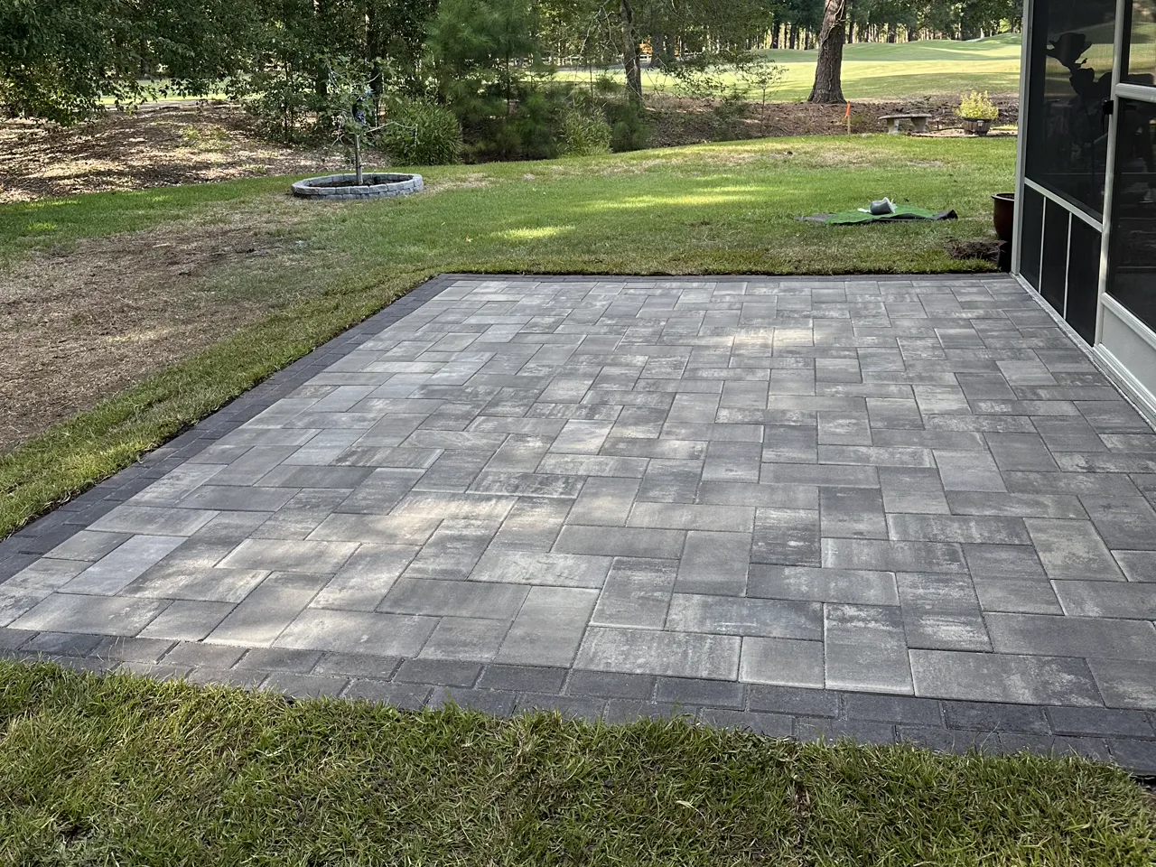 Finished paver patio with a charcoal soldier-course border, fresh sod laid to the edge and a golf course fairway beyond.