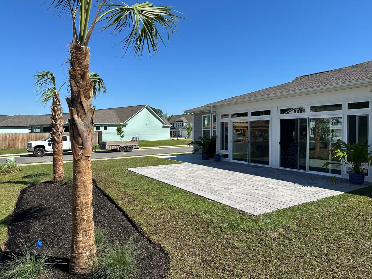 Large paver patio slab across the rear of a home, freshly planted palm alongside.